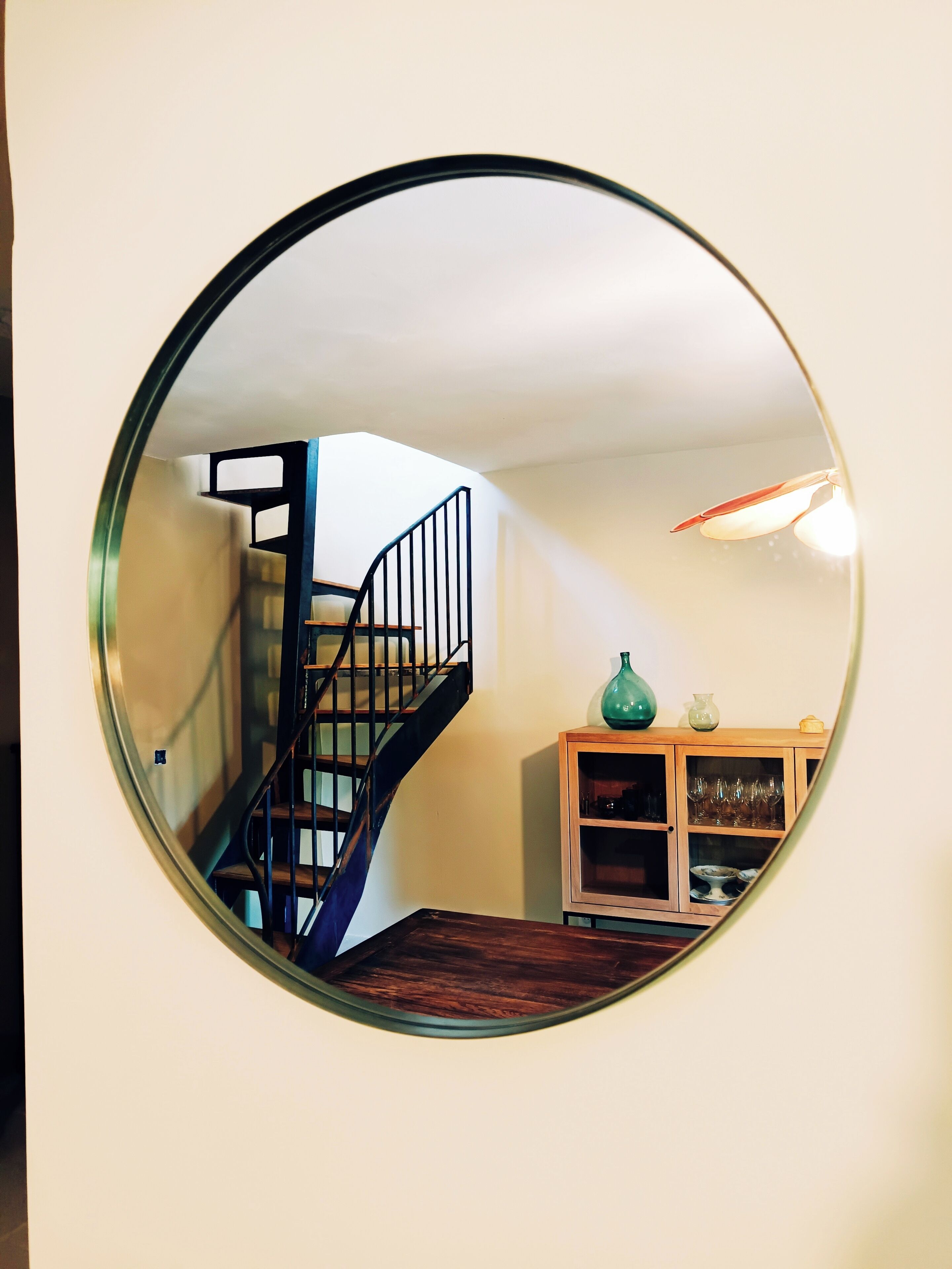 Ein runder Spiegel, der eine Treppe wiederspiegelt, eine Holzkommode mit Gläsern und eine grüne Vase.