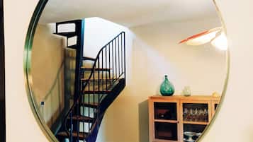 Ein runder Spiegel, der eine Treppe wiederspiegelt, eine Holzkommode mit Gläsern und eine grüne Vase.