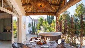 An outdoor dining area with a round table set for two, surrounded by wicker chairs, a hot tub, and a pergola structure.