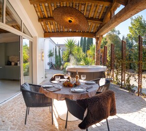 An outdoor dining area with a round table set for two, surrounded by wicker chairs, a hot tub, and a pergola structure.