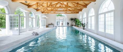 Une grande piscine intérieure à l’eau claire, entourée de fenêtres en arc de cercle et de poutres en bois.
