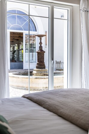 A hotel room with a bed, a view of a courtyard with a fountain, and a building with a large arched window.