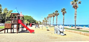 A playground with a red slide and a climbing structure, palm trees, and a beach in the background.