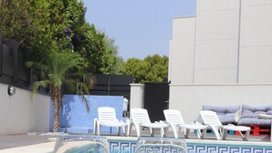 Två glas rött vin med etiketterna ”UPPVÄRMD POOL” och ”VARMVATTENSBAD” står framför en pool med solstolar och en palm i bakgrunden.