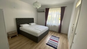 Une chambre à coucher comprenant un lit, une table de chevet, un appareil de climatisation, une fenêtre avec des rideaux et un plancher en bois.
