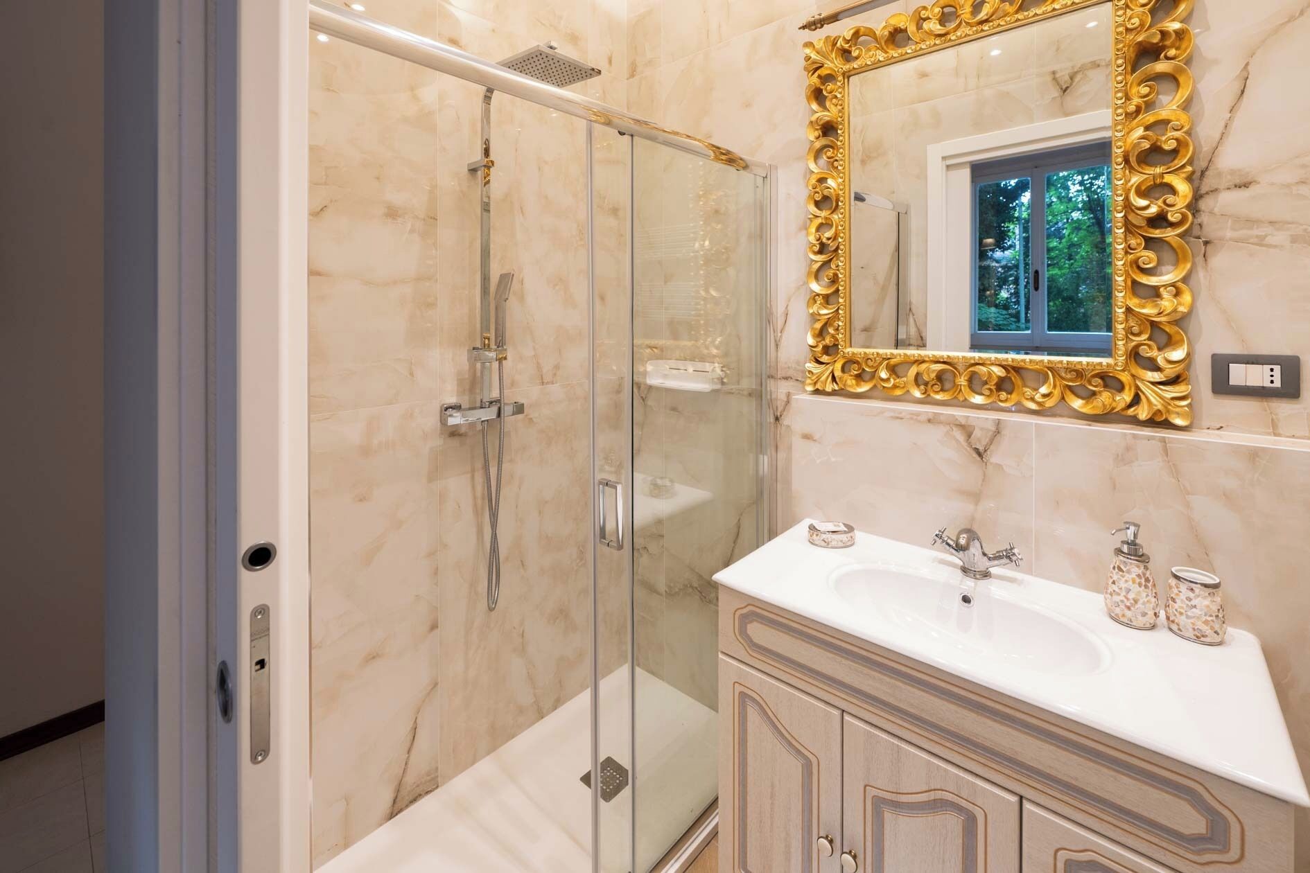 A bathroom with a glass-enclosed shower, a marble vanity with a gold-framed mirror, and a window above the sink.