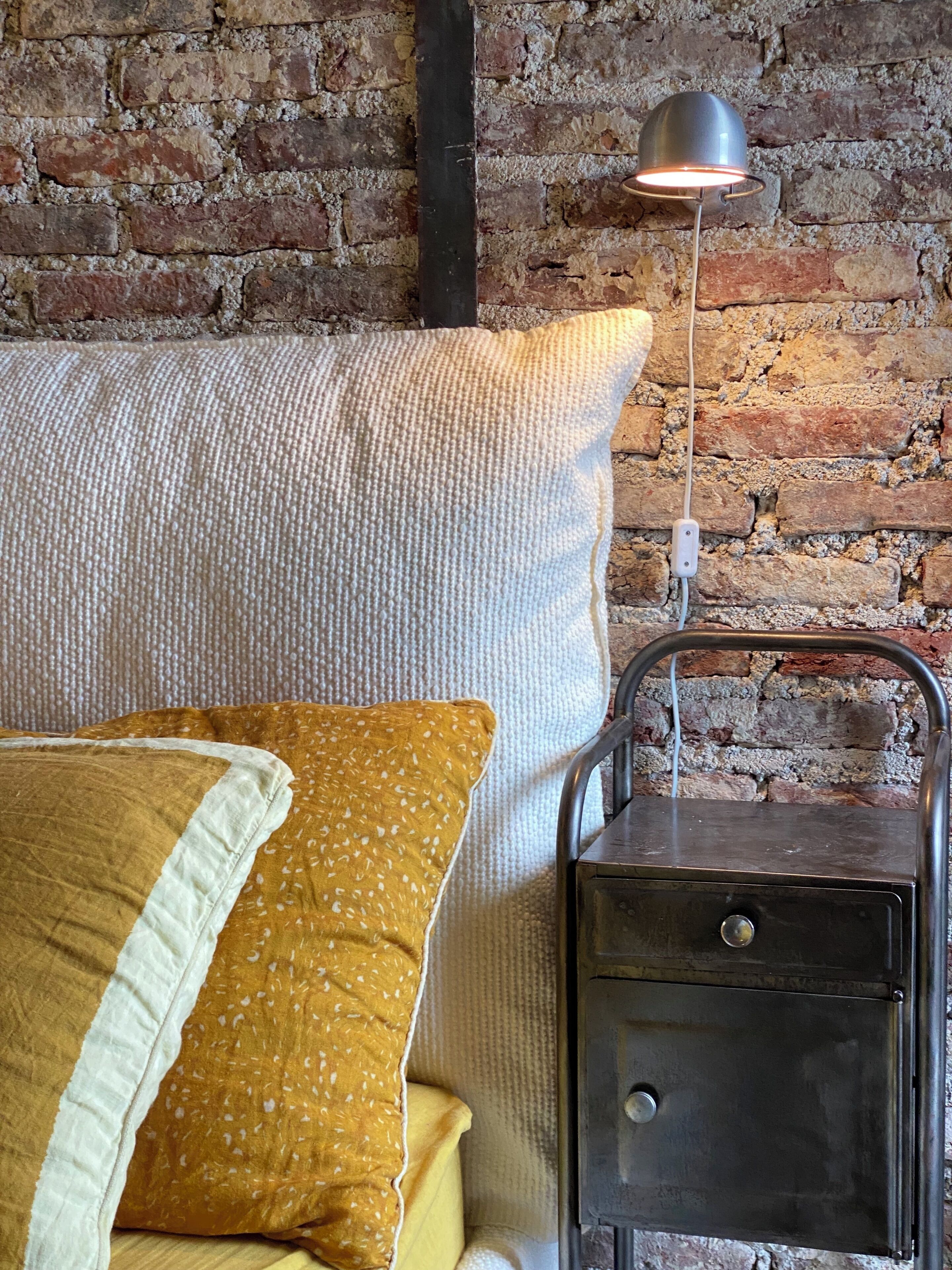 A bed with a white and yellow pillow, a metal nightstand, and a brick wall.