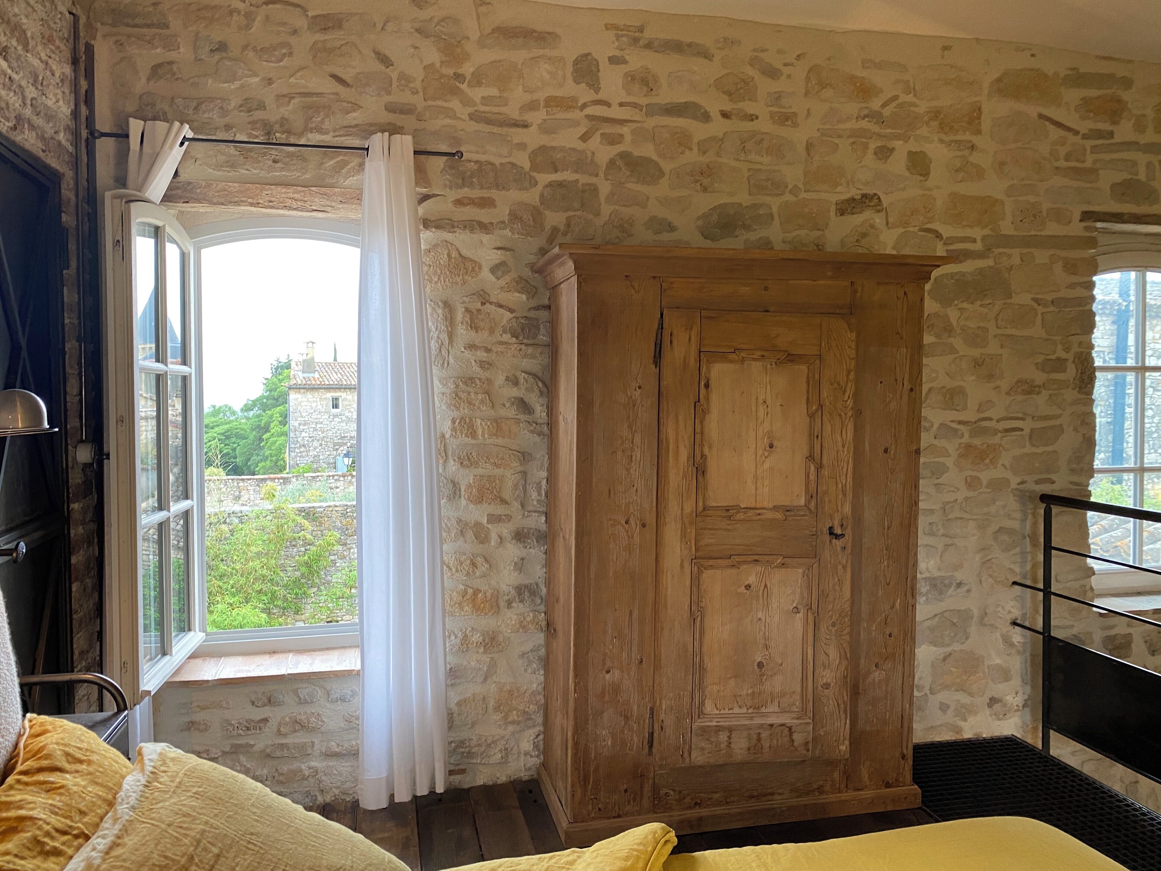 Une pièce aux murs de pierre, avec une armoire en bois, un lit recouvert d’une literie jaune et une fenêtre ornée de rideaux.