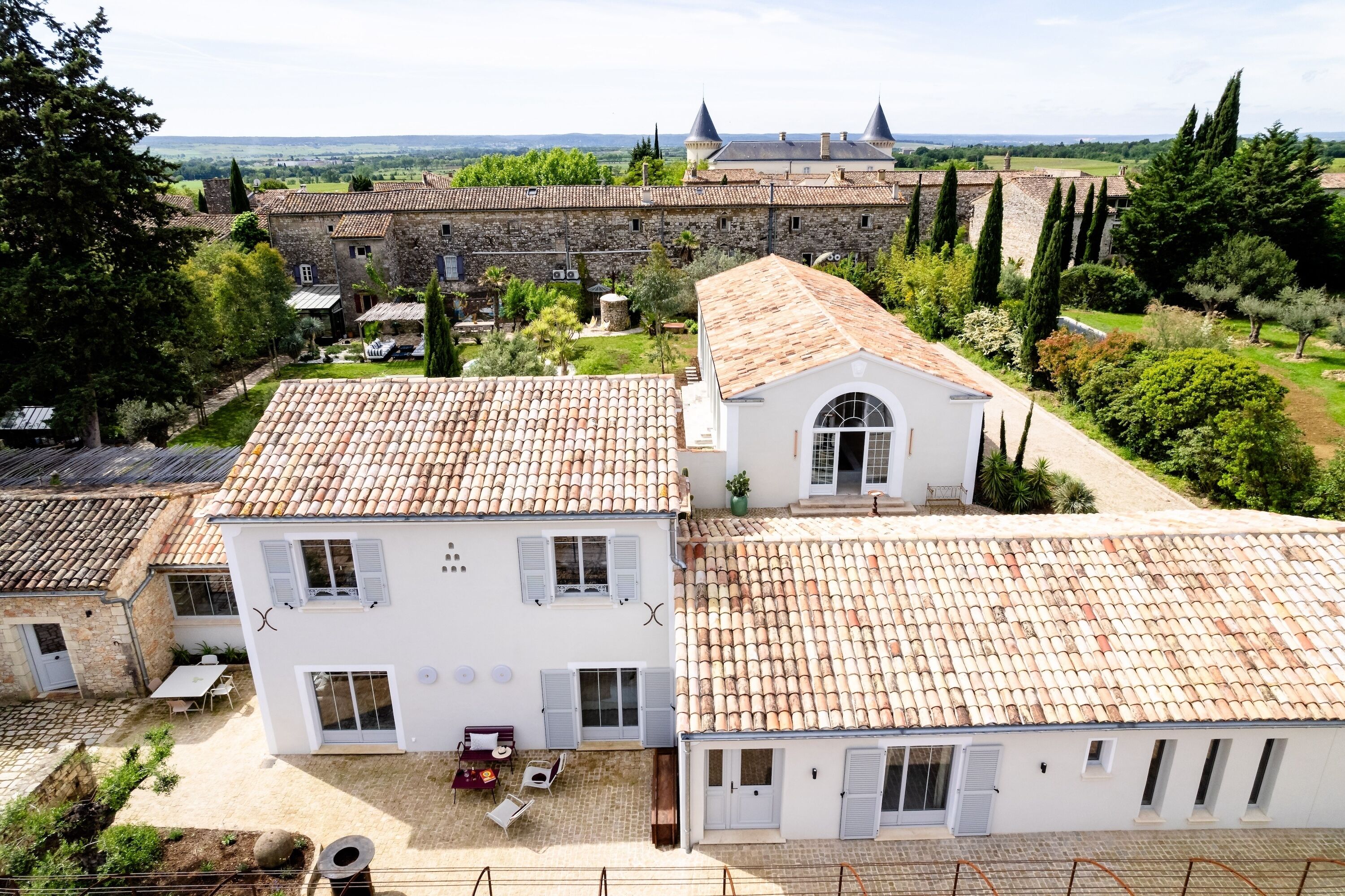 Un bâtiment blanc avec un toit en tuiles, un jardin doté d’une terrasse et une vue sur un château situé au loin.