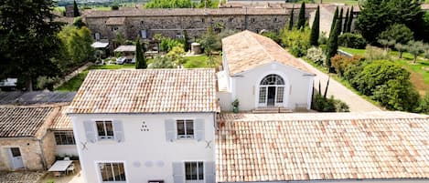 Un bâtiment blanc avec un toit en tuiles, un jardin doté d’une terrasse et une vue sur un château situé au loin.
