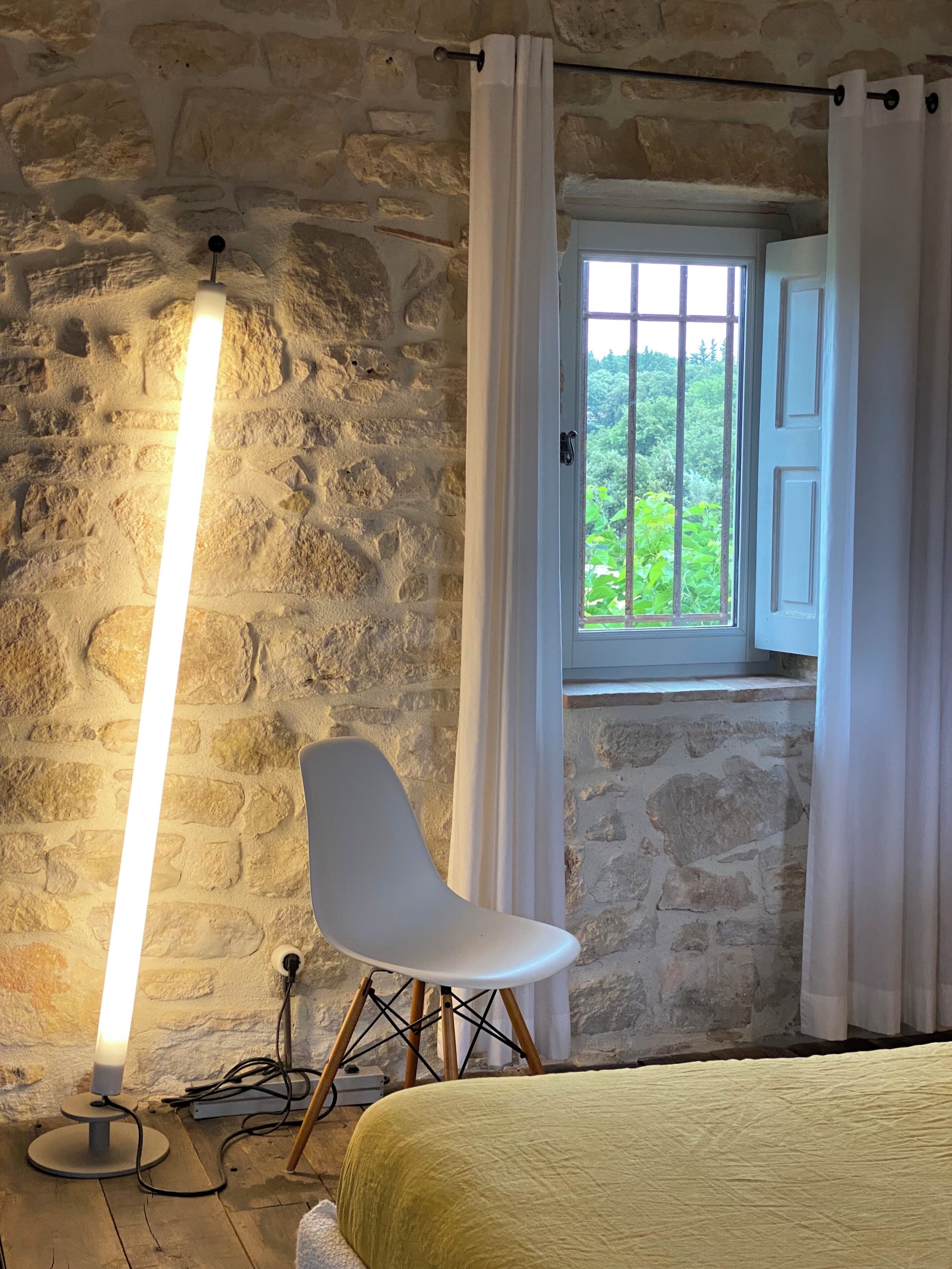 A room with a stone wall, a white chair, a bedside table, a lamp, and a window with curtains.