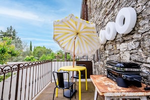 Eine Terrasse mit gelbem Tisch und Stühlen, einem gestreiften Sonnenschirm und einem Grill an einer Steinmauer.