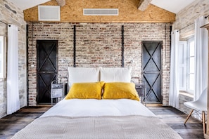 Ein Schlafzimmer mit Backsteinwand, einem Bett mit weißer Bettwäsche und zwei senfgelben Kissen, einem Nachttisch und einem modernen Sessel.