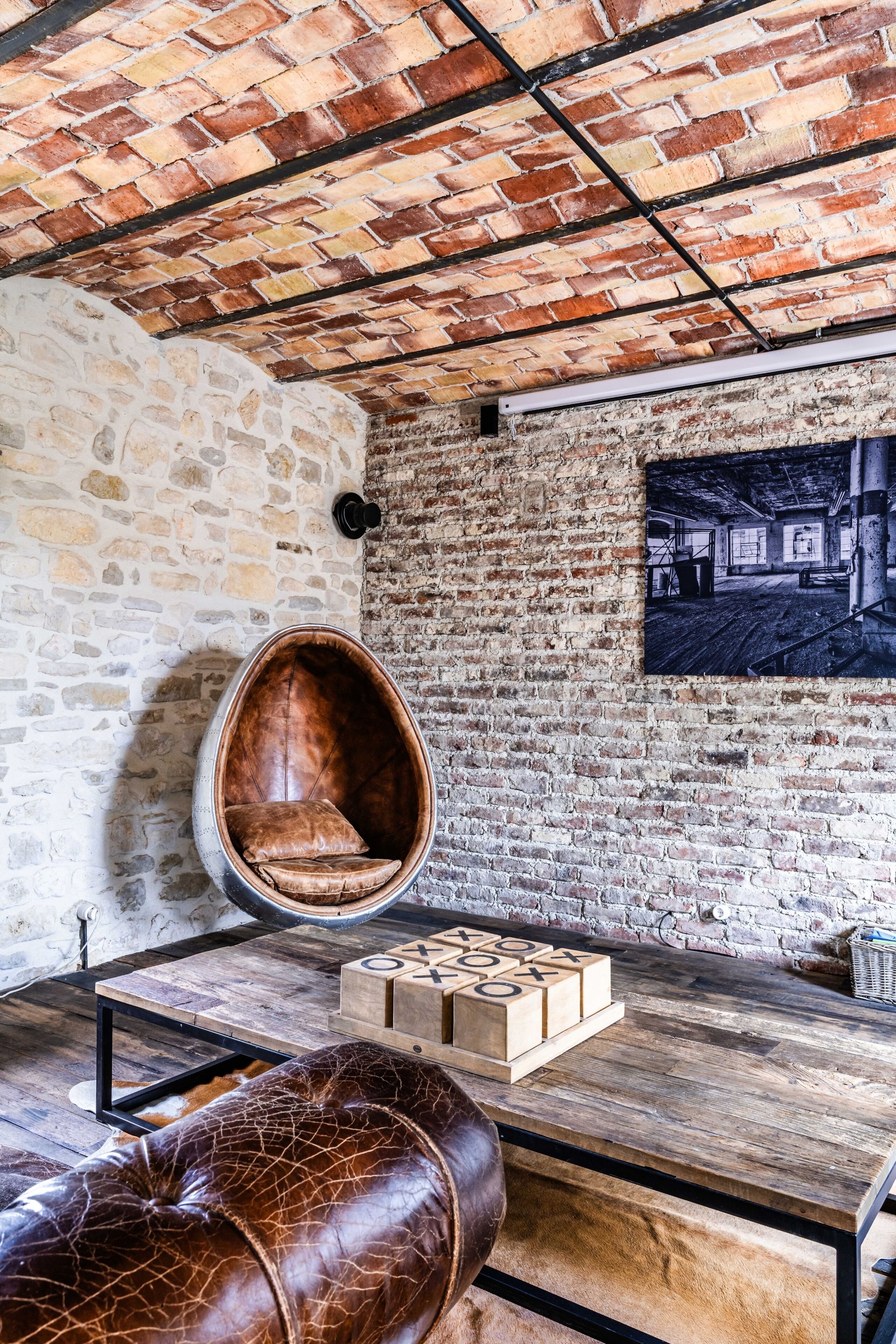 A room with exposed brick walls, a hanging chair, a wooden table with a game, and a leather ottoman.