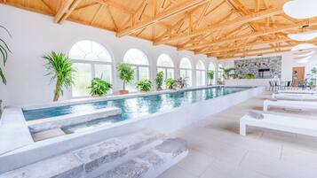 A spacious indoor swimming pool with a high ceiling and arched windows, surrounded by potted plants and a modern kitchen area.