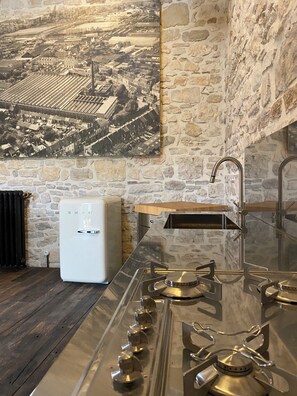 A kitchen with a modern stainless steel stove, a white refrigerator, and a stone wall with a vintage aerial cityscape painting.