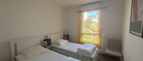 A small, clean room with two single beds, a window with a view of greenery, and a radiator.