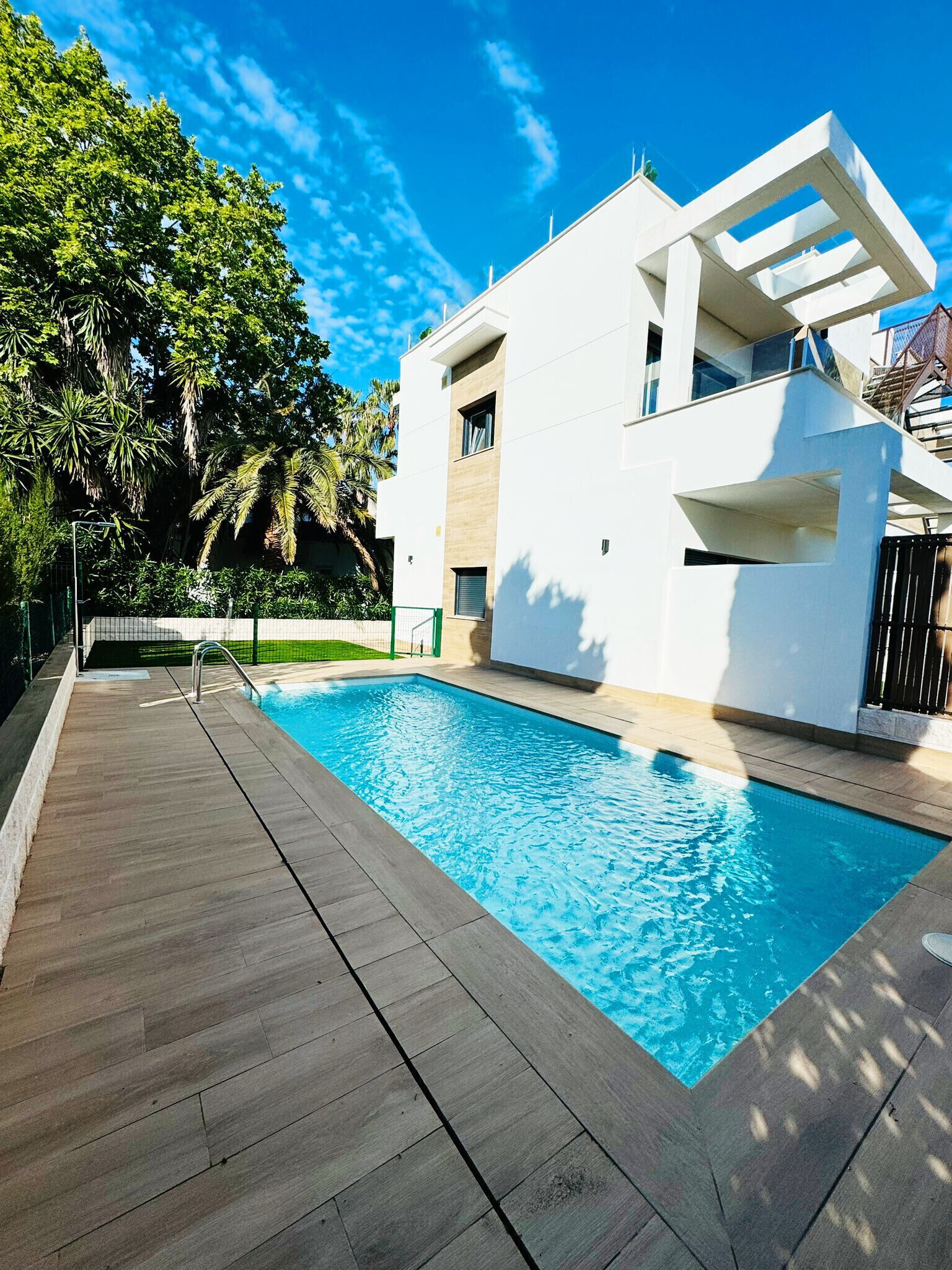 Un área moderna junto a la piscina con una pileta rectangular, terraza de madera y un edificio blanco con balcones.
