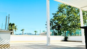 Ein überdachter Außenbereich mit Pergola, Fliesenboden und Blick auf Bäume und entfernte Gebäude.
