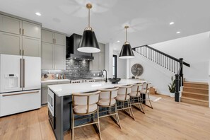 A modern kitchen with a central island, bar stools, a staircase, and a dining area.