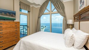 A hotel room with a large bed, a wooden dresser, a nightstand with a lamp, and a view of the ocean.