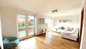 A spacious living room with a large sliding glass door leading to a patio, a white sofa, a blue armchair, and a small table with a lamp. - Happy Waves - Family House - 1 Min to Camber Sands (Camber)