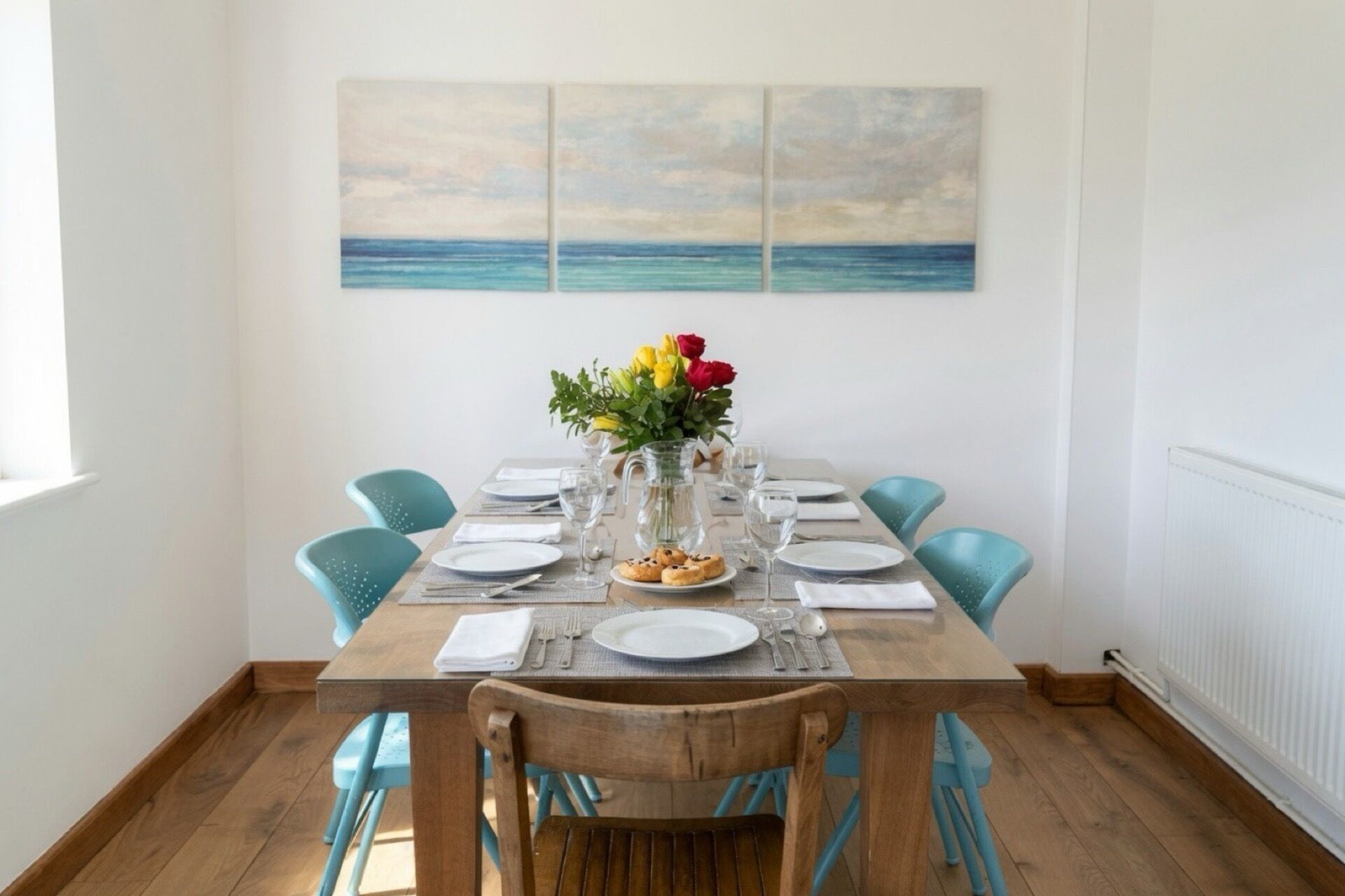 Una mesa de comedor preparada para cuatro con sillas azules, un florero con flores y un cuadro de mar en la pared.
