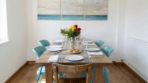 A dining table set for four with blue chairs, a vase with flowers, and a painting of the sea on the wall. - Happy Waves - Family House - 1 Min to Camber Sands (Camber)