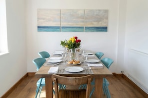 Une table à manger dressée pour quatre personnes, avec des chaises bleues, un vase rempli de fleurs et un tableau représentant la mer accroché au mur.