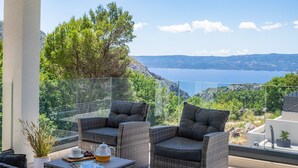 Un balcon doté de meubles en osier, d’une rambarde en verre et offrant une vue sur un paysage côtier.