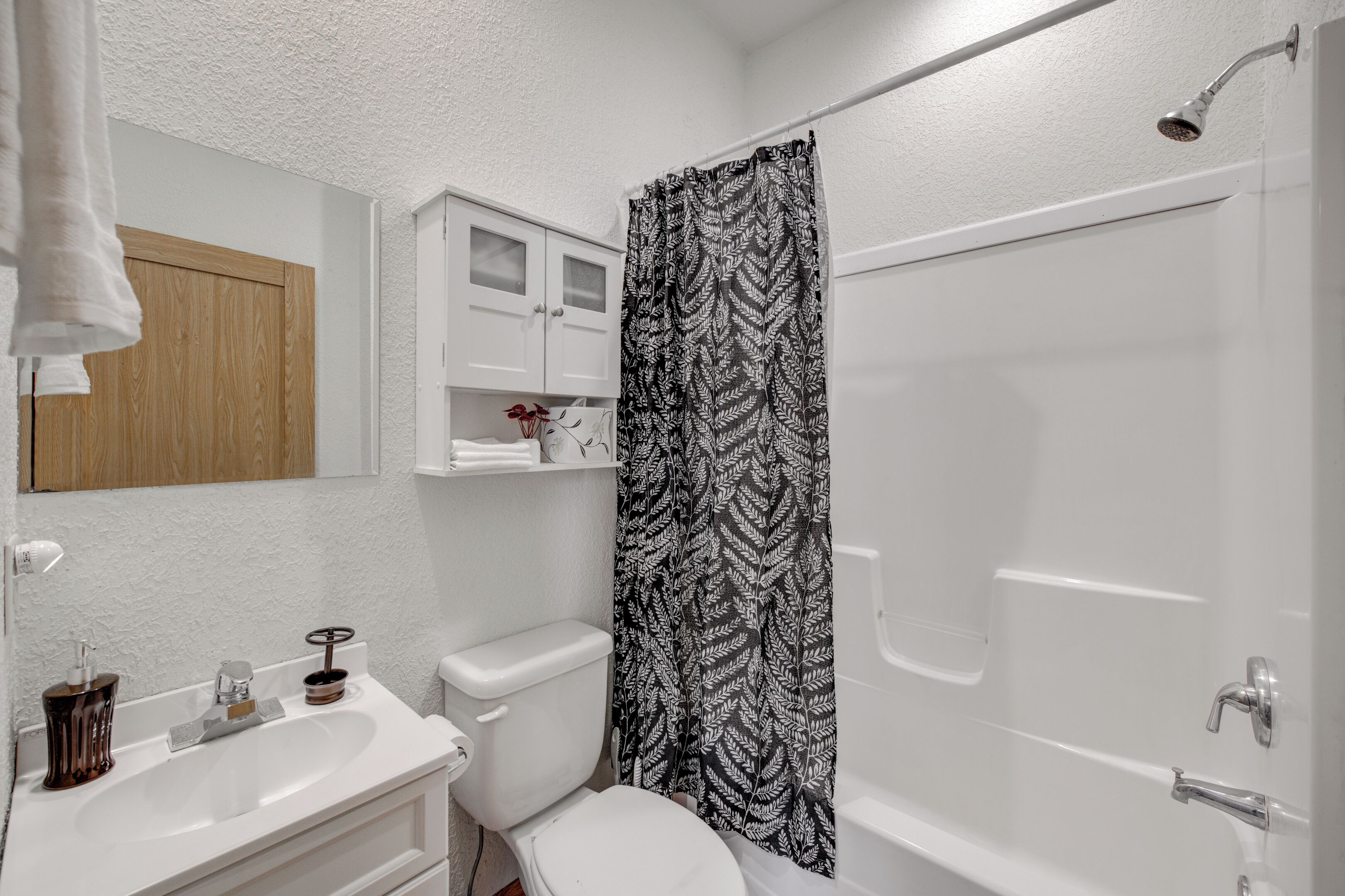 Un baño con lavamanos, inodoro y bañera blancos. Hay una cortina de ducha con estampado de hojas, una puerta de madera y un mueble blanco sobre el lavamanos.