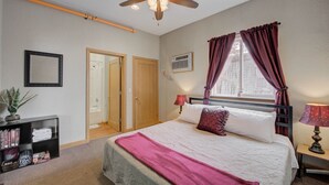 A bedroom with a bed, a ceiling fan, a window with curtains, a bedside lamp, a bookshelf, and a door. - Three Connected Quirky Condos By Beach! - Twilight Condo (Pepin)
