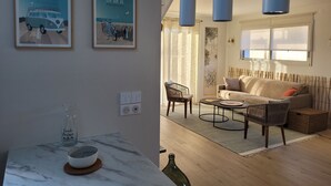 A living room with a sofa, a coffee table, and a dining area with a marble table. There are two framed pictures on the wall, a hanging light fixture, and a window with blinds. - Maison de Vacances à 2 pas de la Plage et des Commerces (Pornic)