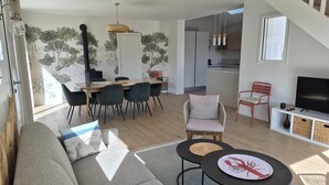 A modern living room with a sofa, a dining area with chairs, a TV, and a kitchen in the background. - Maison de Vacances à 2 pas de la Plage et des Commerces (Pornic)