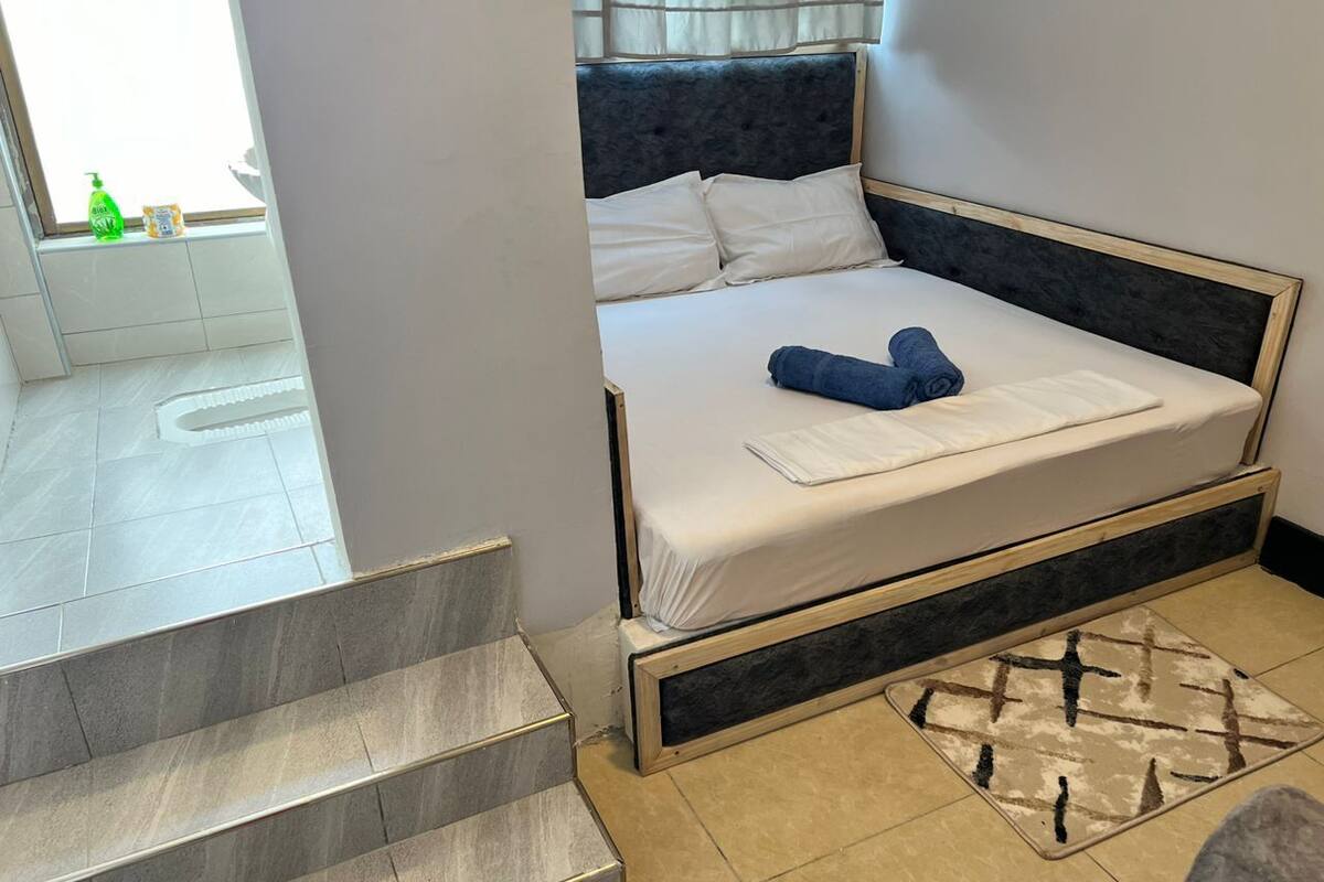 Un lit avec une literie blanche et un oreiller bleu, un tapis à motifs et une fenêtre avec des rideaux.