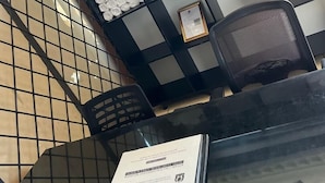 Egy előtér, melyben egy üveglapos asztal található, rajta egy oklevél, egy fekete szék, és a falon egy rácsmintázat és feltekert törölközők láthatók.