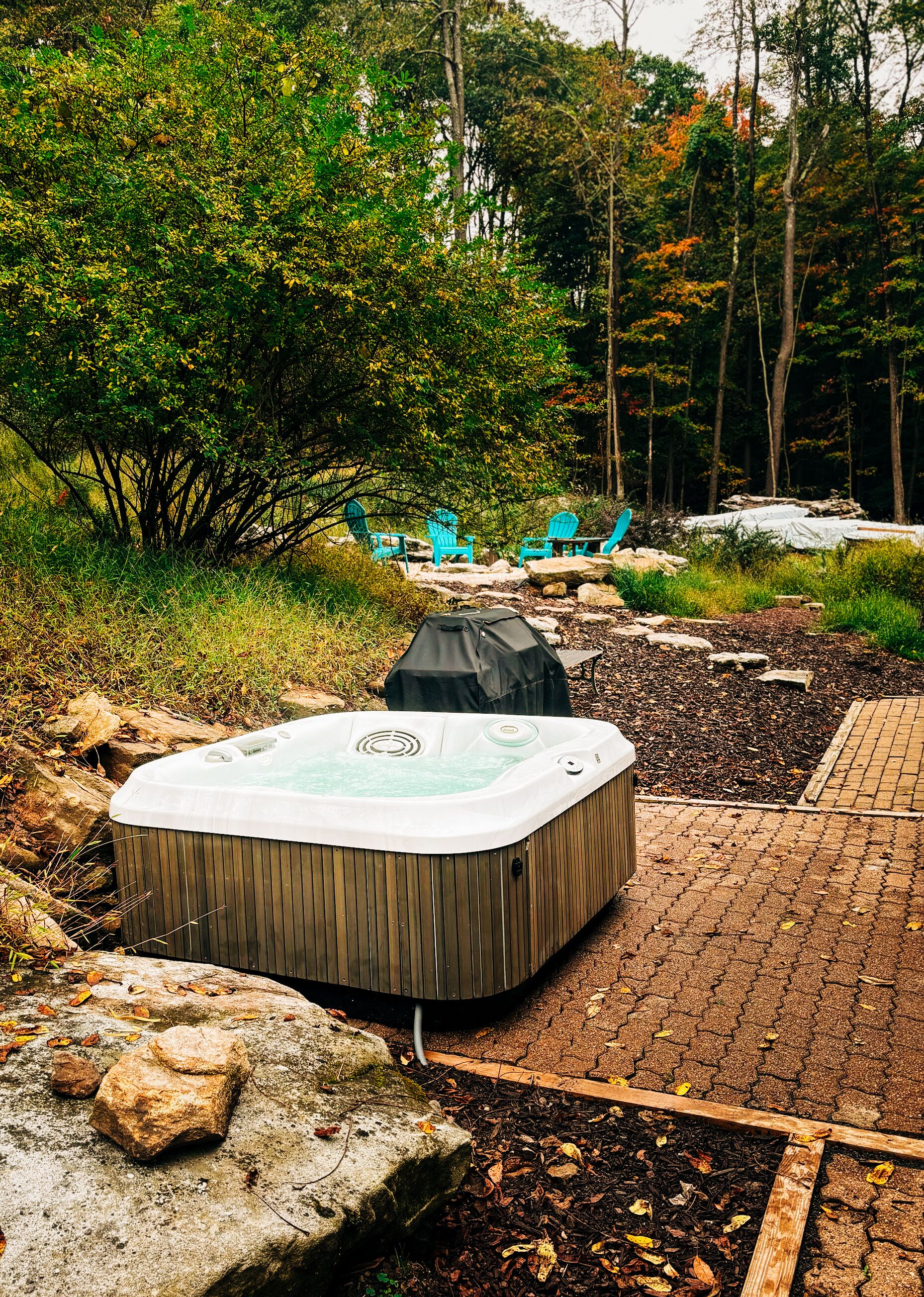 A hot tub surrounded by rocks and a brick pathway, with blue chairs and a grill in the background.
