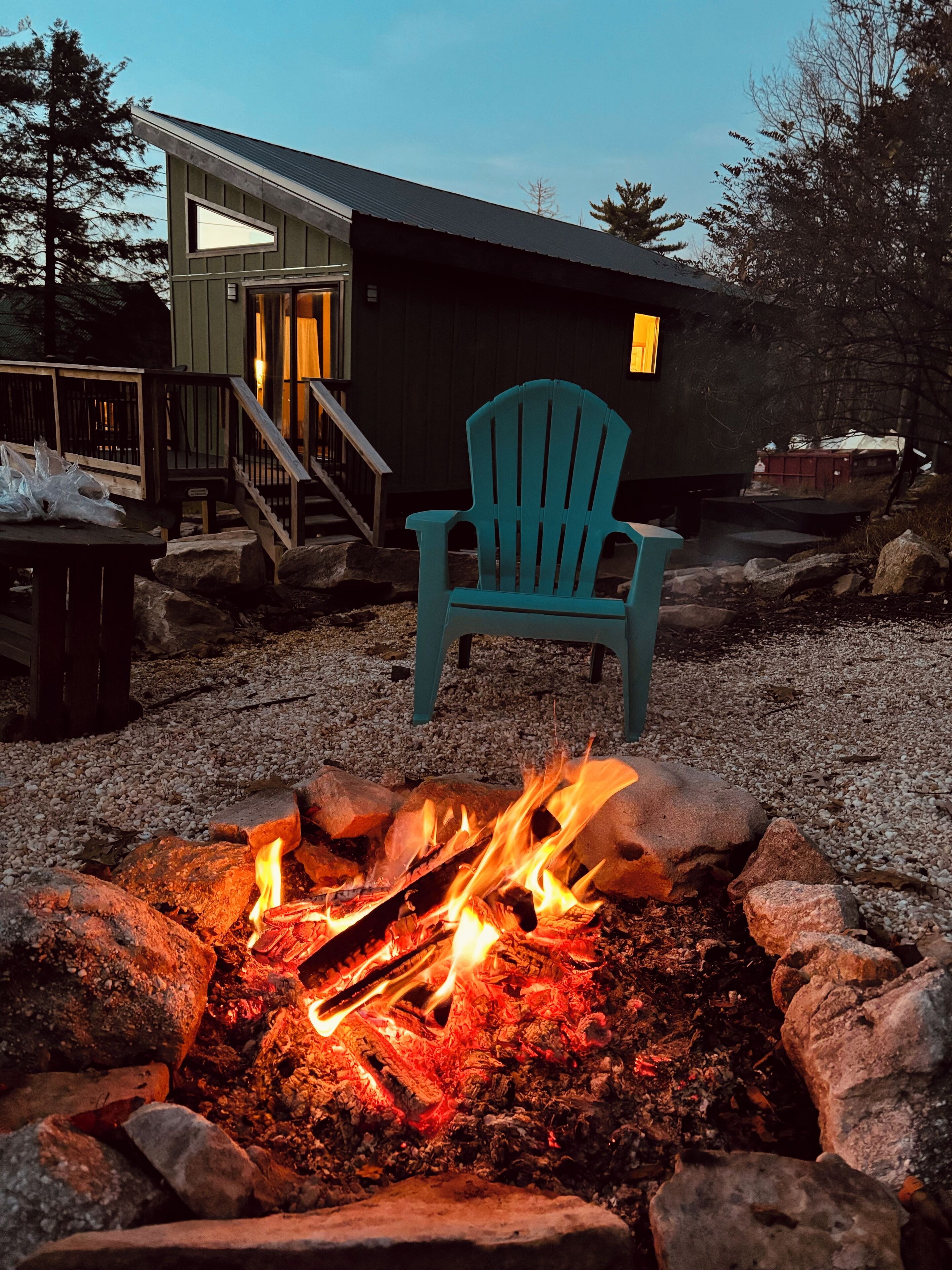 Un rincón exterior acogedor con una chimenea, una silla Adirondack turquesa y una terraza de madera que conduce a una casa con puerta de vidrio.