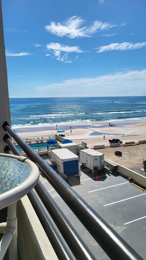 A balcony with a view of the beach and ocean. There are lounge chairs and umbrellas on the beach, and a parking area with a few vehicles. - Spectacular, Oceanview Condo, Pool, Hot Tub, Gorgeous Views sunrise to sunset! (Daytona Beach Shores)