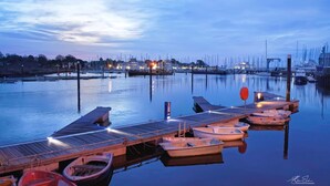 A marina with several boats docked at wooden piers, calm water, and a cityscape in the background during twilight. - Newgate Cottage by NewFo rest (Lymington)