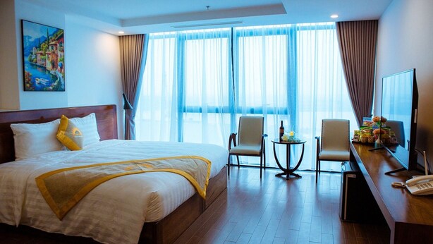 A hotel room with a large bed, two armchairs, a small table, a desk with a computer and phone, and a large window with curtains. - Solis Hotel (Hai Phong)