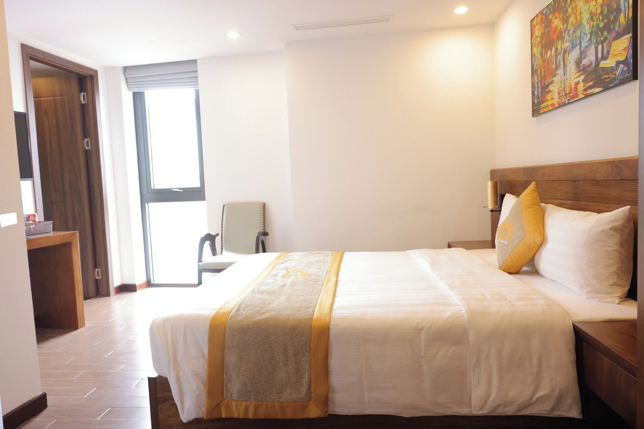A hotel room with a large bed, a wooden headboard, a painting on the wall, and a window with curtains.