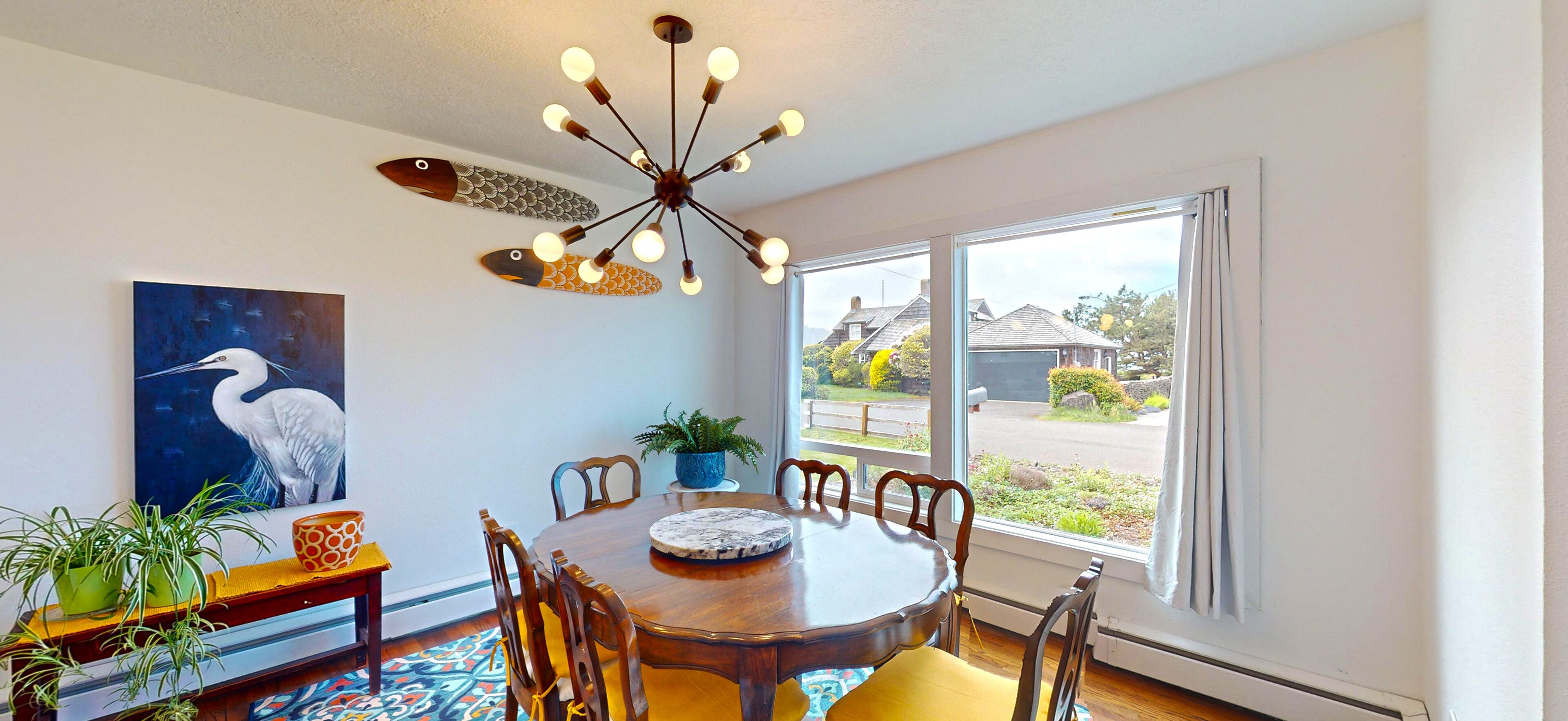 A dining area with a round table, chairs, a ceiling light fixture, and a large window with a view of houses.