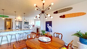 A modern kitchen with a central island, bar stools, a dining table with chairs, and a ceiling light fixture.