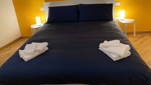 Una cama bien hecha con ropa de cama azul oscuro, dos toallas blancas dobladas al pie de la cama y dos lámparas de noche en mesitas blancas.