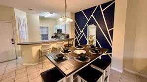 Una cocina moderna con armarios blancos, microondas y estufa. Hay una zona de comedor con una mesa de madera, cuatro sillas y un espejo decorativo en la pared.