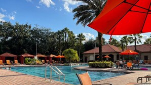 Un área de piscina con sillones de descanso y sombrillas, rodeada de palmeras y un edificio con tejado de tejas.