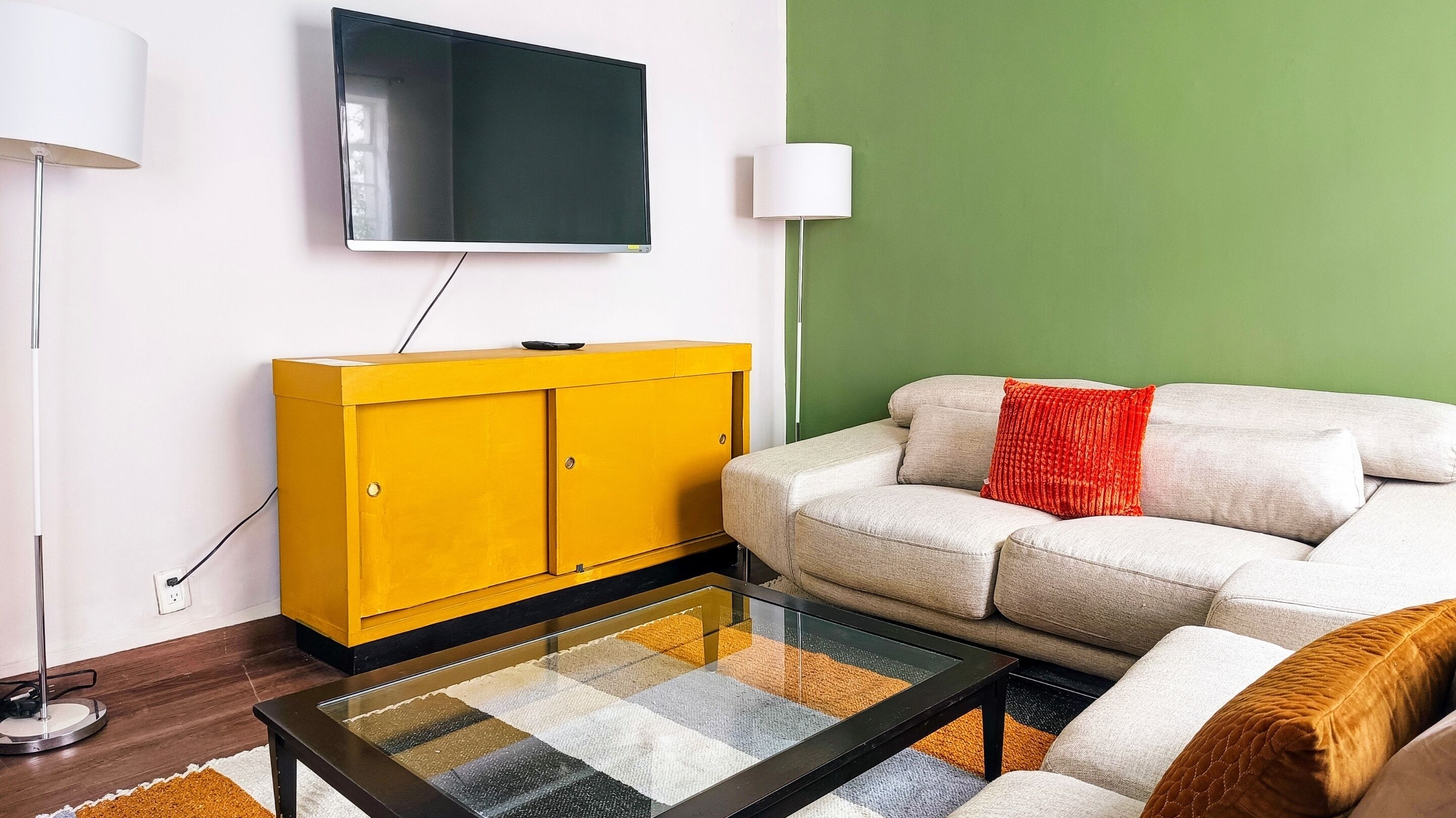 Una sala de estar con un sofá, un armario amarillo, una mesa de centro de vidrio y un televisor en la pared.