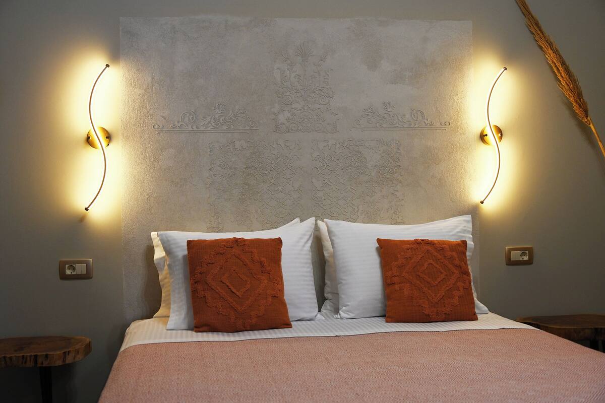 A neatly made bed with white and brown pillows, a pink blanket, and two wall-mounted light fixtures on either side.