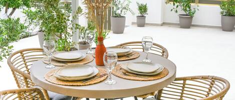 A round table set for four with matching plates, cutlery, and wine glasses, surrounded by wicker chairs.